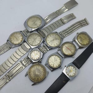 Puede incluir: Una colección de relojes de pulsera vintage con correas de metal plateado y esferas. Los relojes tienen varios diseños de esfera, incluyendo formas redondas, cuadradas y rectangulares. Algunos relojes tienen correas de cuero negro.