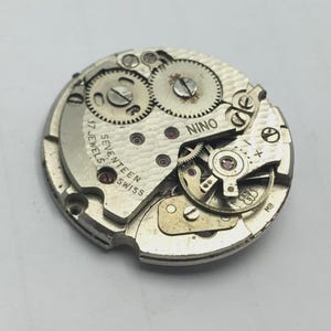 May include: A vintage watch movement with visible gears, screws, and the text "NINO" and "SEVENTEEN SWISS JEWELS". The metal components are silver-toned, with red jewels. The watch mechanism is circular.