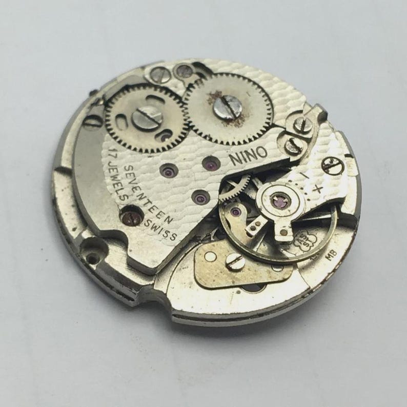 May include: A vintage watch movement with visible gears, screws, and the text "NINO" and "SEVENTEEN SWISS JEWELS". The metal components are silver-toned, with red jewels. The watch mechanism is circular.