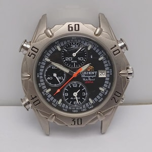 May include: Silver-toned Orient chronograph watch with a black dial. The watch features multiple sub-dials, a date display, and luminous hands. The bezel has markings for timekeeping, and the watch is water-resistant to 100m.