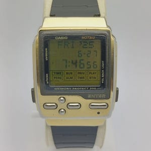 May include: A vintage Casio Hotbiz digital watch with a gold-tone case and a black strap. The watch face displays the date and time. The watch has several buttons and function labels, including "ENTER" and "MEMORY PROTECT 200".