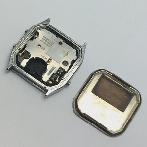 May include: An open, silver-toned watch case with the back cover detached, revealing the internal components. The watch face has text that reads "CASIO-JAPAN" and other markings. The back cover is a silver frame with a brown interior.