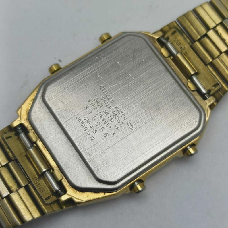 May include: A gold-coloured Citizen watch with a rectangular face and a matching metal strap. The watch's back is visible, displaying text including "CITIZEN WATCH CO." and "WATER RESIST." The strap is made of small, connected links.