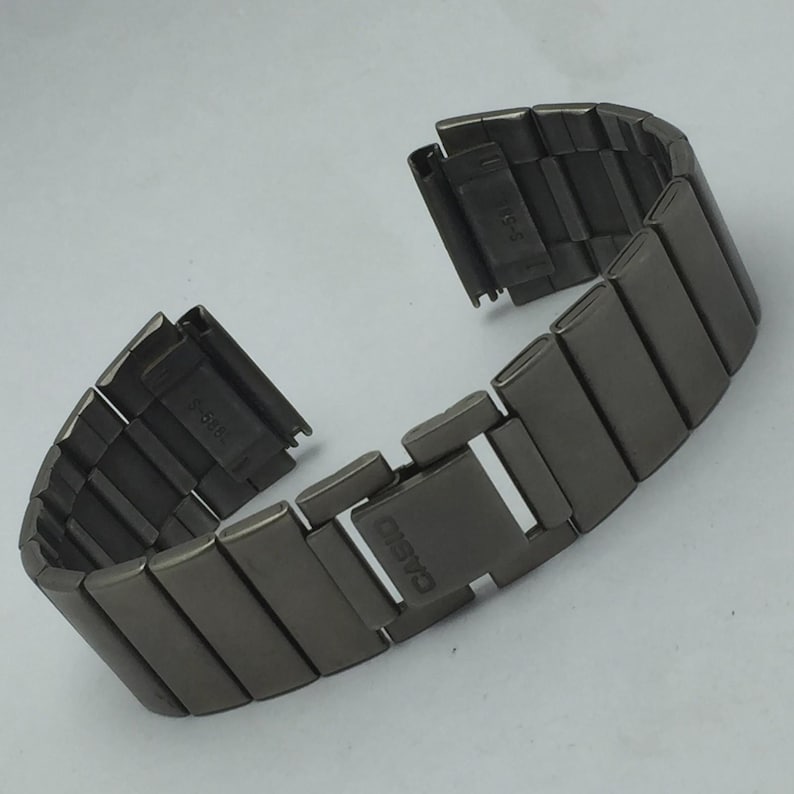 Puede incluir: Una correa de reloj de metal gris oscuro con eslabones rectangulares. La correa est&aacute; desabrochada, mostrando la marca Casio en el cierre. La correa est&aacute; dise&ntilde;ada para un reloj.