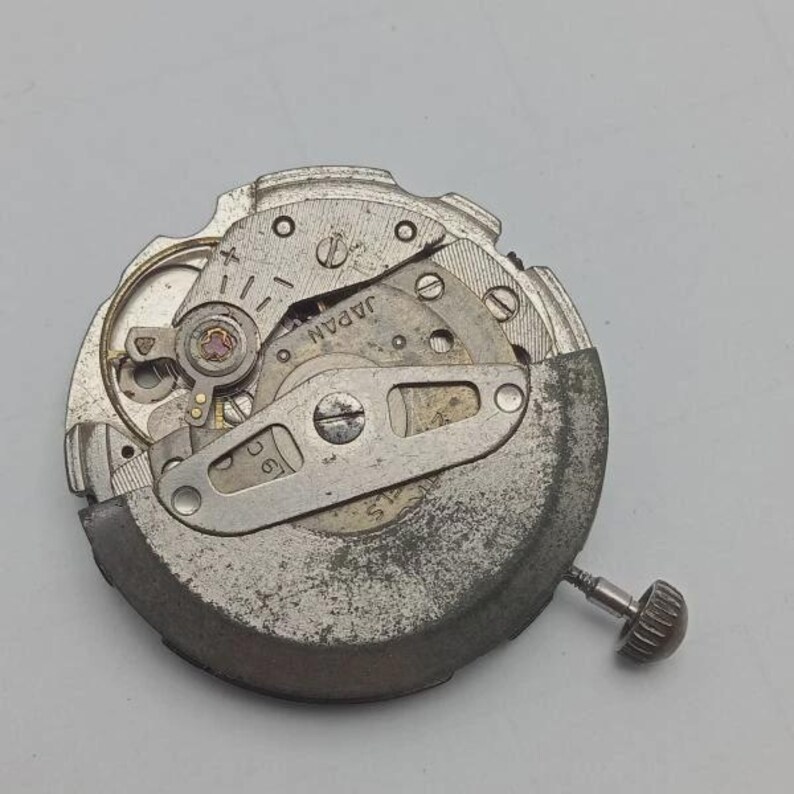 Peut inclure: Gros plan d'un mouvement de montre m&eacute;canique avec des engrenages, des vis et le mot "JAPAN" visible. Les composants m&eacute;talliques sont argent&eacute;s avec une texture. Une petite couronne est pr&eacute;sente.