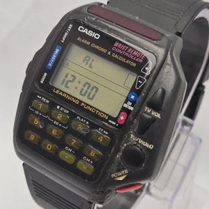 Puede incluir: Un reloj Casio Wrist Remote Controller vintage, negro, con un teclado de calculadora y una pantalla LCD que muestra "12:00". Incluye alarma, crono y funci&oacute;n de aprendizaje. El reloj tiene una correa negra y varios botones de control.