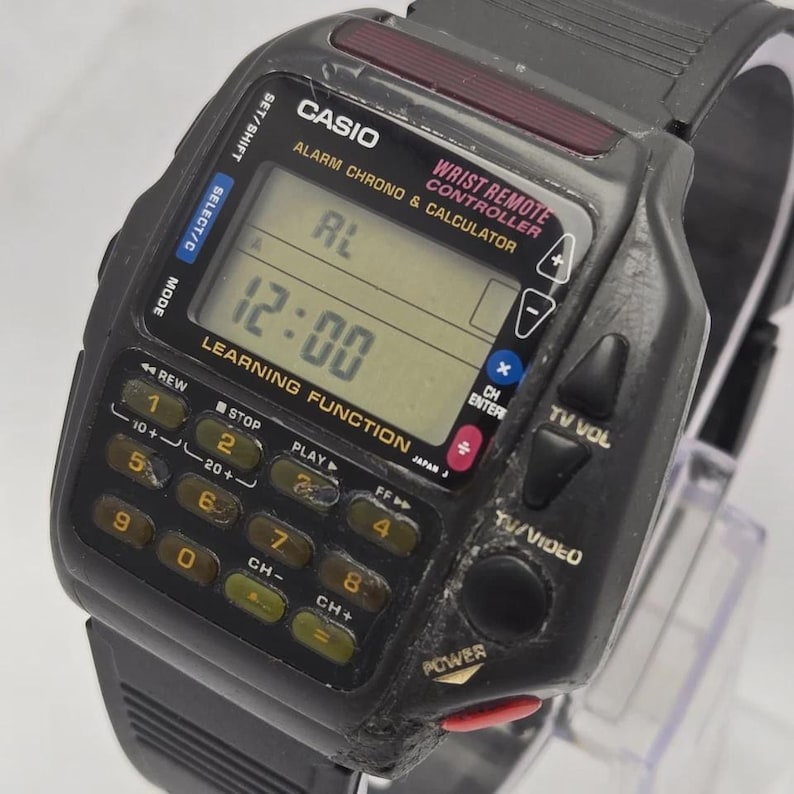 Puede incluir: Un reloj Casio Wrist Remote Controller vintage, negro, con un teclado de calculadora y una pantalla LCD que muestra "12:00". Incluye alarma, crono y funci&oacute;n de aprendizaje. El reloj tiene una correa negra y varios botones de control.