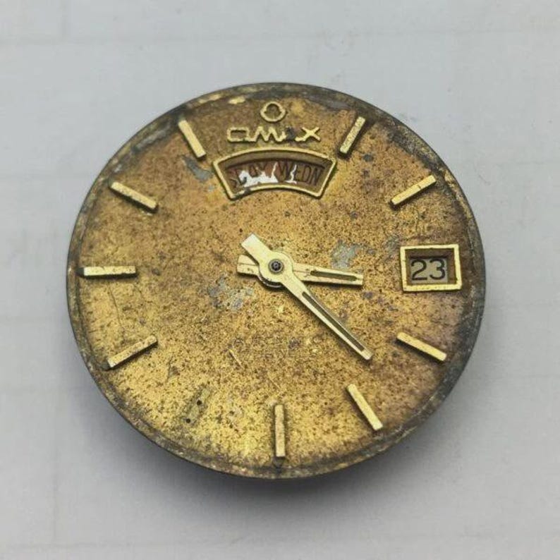 May include: Aged gold-toned watch face with a day-date display and the number 23. The face has gold hands and hour markers. The brand name "AMX" is visible above the day-date window. The watch face has a worn, vintage appearance.