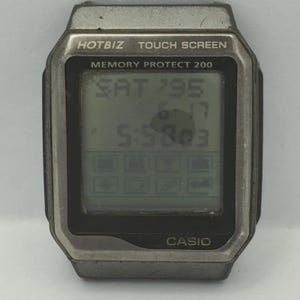 Puede incluir: Un reloj táctil Hotbiz plateado y negro. La pantalla digital muestra la fecha, la hora y varios iconos. La esfera del reloj dice "MEMORY PROTECT 200" y "CASIO".