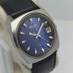 May include: A vintage Seiko automatic wristwatch with a blue dial and a black leather strap. The watch has a silver-toned case and features a date display. The dial is marked with the Seiko logo and "17 Jewels".
