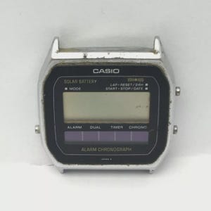 May include: A silver Casio digital watch face with a black display. The watch face has the text "CASIO" at the top, with "SOLAR BATTERY" and "ALARM CHRONOGRAPH" below. The display shows various functions including alarm, dual, timer, and chrono.