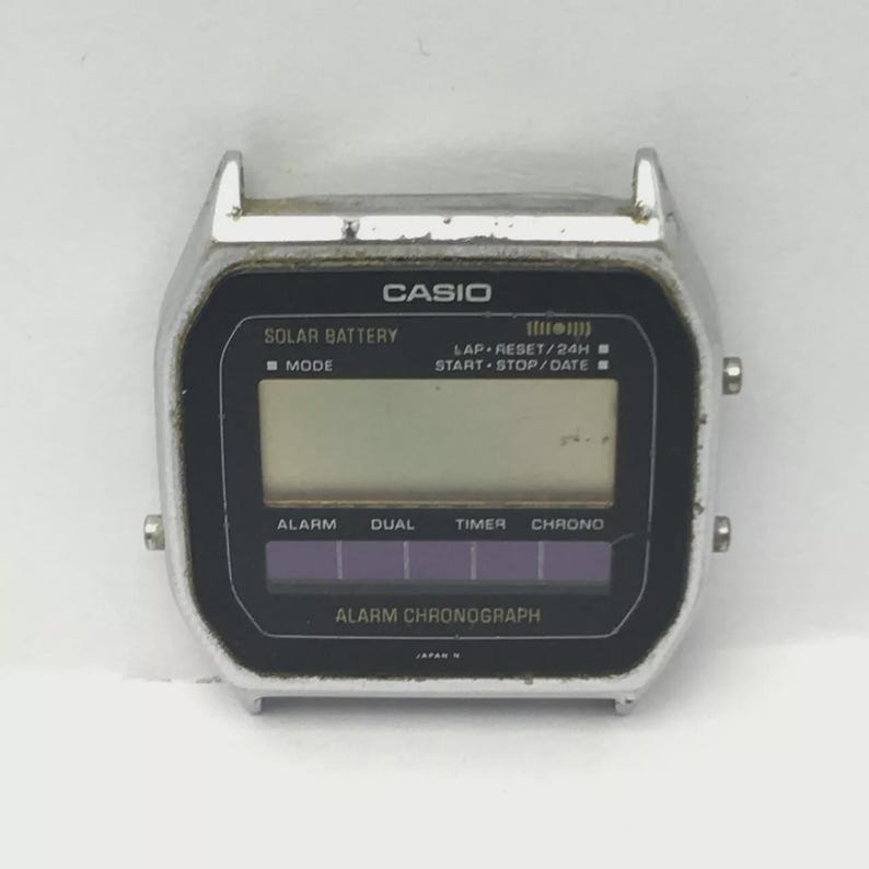 May include: A silver Casio digital watch face with a black display. The watch face has the text "CASIO" at the top, with "SOLAR BATTERY" and "ALARM CHRONOGRAPH" below. The display shows various functions including alarm, dual, timer, and chrono.