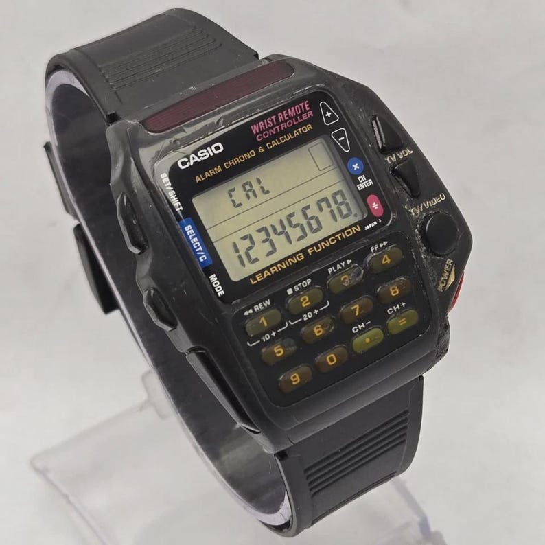 Puede incluir: Reloj de pulsera Casio negro con funciones de calculadora y mando a distancia. La pantalla muestra el n&uacute;mero 12345678. Tiene botones para el volumen de la televisi&oacute;n, la selecci&oacute;n de canales y otras funciones.