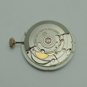 May include: A close-up of a watch movement with intricate gears and components. The watch mechanism is silver and gold, with the text "TWENTY-FIVE 25 JEWELS SWISS MADE" visible. A gold crown extends from the side.