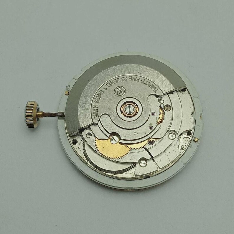 May include: A close-up of a watch movement with intricate gears and components. The watch mechanism is silver and gold, with the text "TWENTY-FIVE 25 JEWELS SWISS MADE" visible. A gold crown extends from the side.