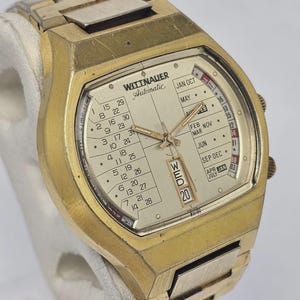May include: Vintage gold-toned Wittnauer Automatic watch with a rectangular face. The watch features a calendar display with the day and date, and a gold-toned metal band. The watch face displays the day of the week and the date.