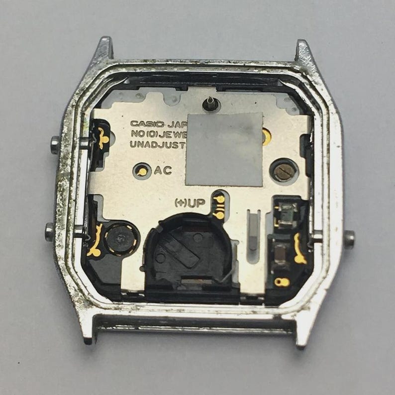 May include: A silver-toned watch case with the internal components visible. The watch face includes the text "CASIO JAPAN NO(0)JEWELS UNADJUST". The internal components are silver and gold-toned, with a black battery compartment.