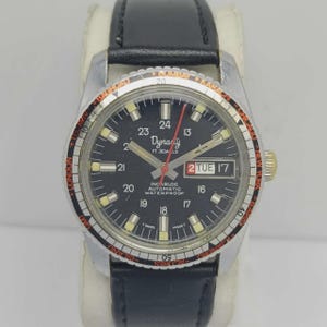Op de afbeelding: Een vintage Dynasty polshorloge met een zwarte wijzerplaat, dag- en datumweergave. Het horloge heeft een zilverkleurige kast, een zwarte leren band en een draaibare bezel met wereldsteden. De wijzerplaat toont de tekst "Dynasty 17 Jewels".
