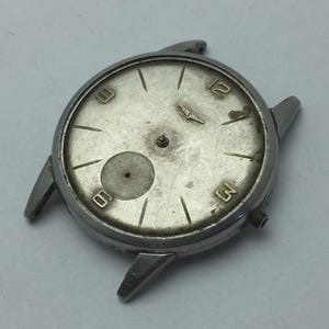 May include: A vintage silver-toned watch face with a worn, textured appearance. The watch face features black numerals at the 3, 6, and 12 o'clock positions, with a small logo above the 12. The watch case has lugs for attaching a strap.