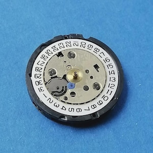 May include: A watch movement with a white dial featuring black numerals from 1 to 31, indicating the days of the month. The movement has a silver-colored center with gears and a gold-colored central gear. The outer ring is black.