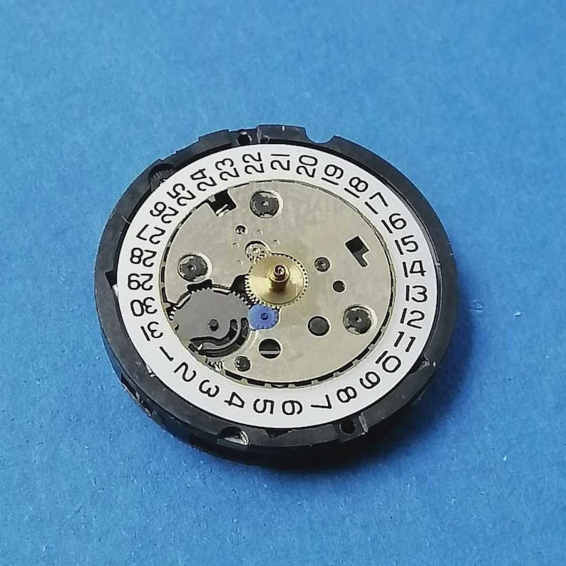 May include: A watch movement with a white dial featuring black numerals from 1 to 31, indicating the days of the month. The movement has a silver-colored center with gears and a gold-colored central gear. The outer ring is black.