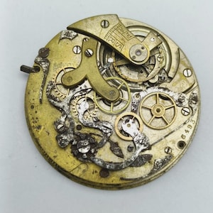 May include: Close-up of a vintage watch movement with intricate gears, springs, and levers. The metal components are in shades of gold and silver, showing signs of age and wear. The number "6493" is visible.