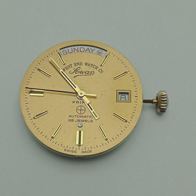 May include: A gold-toned watch face with the text "SUNDAY" at the top. The watch face has gold hands, a date window, and the words "WEST END WATCH CO" and "Sowan" are visible. The watch is marked "SWISS MADE".