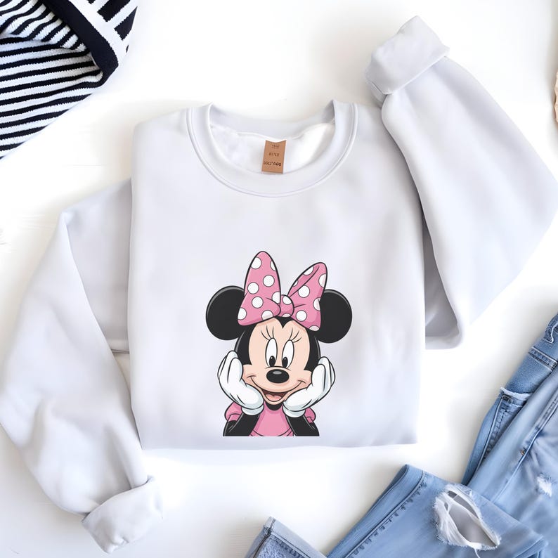 May include: White sweatshirt with a Minnie Mouse graphic. The graphic shows Minnie Mouse with a pink bow with white polka dots, black ears, and a pink dress. The sweatshirt is folded with blue jeans visible.
