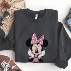 May include: A dark grey sweatshirt with a Minnie Mouse graphic. The design shows Minnie Mouse with a pink bow with white polka dots, a pink dress, and white gloves. The sweatshirt is folded on a white surface.
