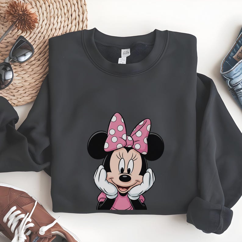 May include: A dark grey sweatshirt with a Minnie Mouse graphic. The design shows Minnie Mouse with a pink bow with white polka dots, a pink dress, and white gloves. The sweatshirt is folded on a white surface.