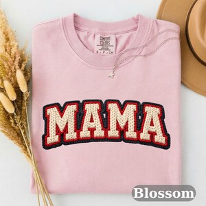 May include: A light pink Comfort Colors t-shirt with the word "MAMA" in a cream-colored, textured font with red and black trim. A silver necklace with a star pendant and a brown hat are visible. The word "Blossom" is at the bottom.