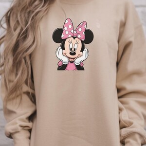 May include: A beige sweatshirt with a Minnie Mouse graphic. The graphic depicts Minnie Mouse with a pink bow with white polka dots, a pink dress, and white gloves. Minnie is resting her chin on her hands.
