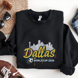 May include: Black sweatshirt featuring a Dallas skyline graphic with the text "Dallas World Cup 2026" in yellow and blue. The design includes a soccer ball and colorful paint splatter accents, perfect for soccer fans.