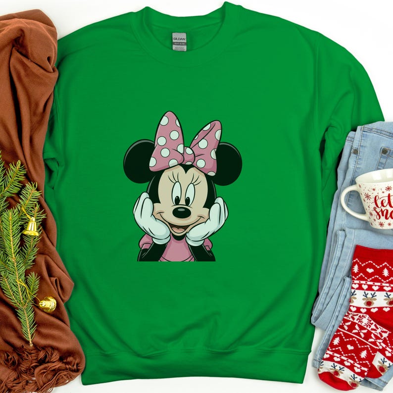 May include: Green sweatshirt with a Minnie Mouse graphic. Minnie Mouse is shown with a pink bow with white spots, a pink dress, and white gloves. The sweatshirt is next to a brown cloth, a pair of jeans, a mug, and a pair of red socks.