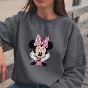 May include: A dark grey sweatshirt with a Minnie Mouse graphic. The graphic shows Minnie Mouse with a pink polka-dot bow, a pink dress, and white gloves. The design is centrally placed on the sweatshirt.