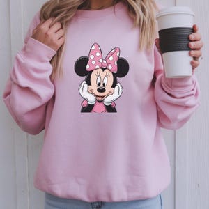 May include: A light pink sweatshirt with a Minnie Mouse graphic. The graphic shows Minnie Mouse with a pink bow with white polka dots. The sweatshirt is a solid light pink colour.