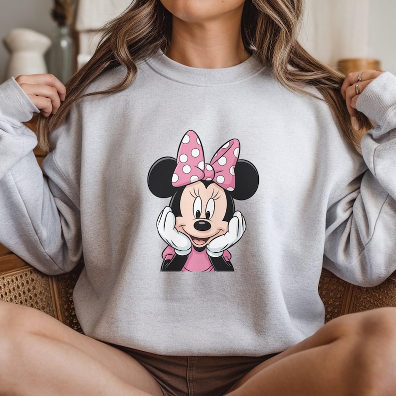 May include: A light grey sweatshirt with a Minnie Mouse graphic. The graphic depicts Minnie Mouse with a pink bow with white polka dots, a pink dress, and white gloves. The sweatshirt is a casual style.