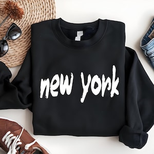 May include: Black crew neck sweatshirt with the white text "new york". The sweatshirt is folded on a white surface, with sunglasses, a woven basket, and brown shoes in the background. The sweatshirt is made of a soft material.
