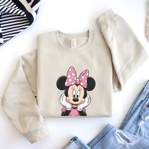 May include: A beige sweatshirt featuring a Minnie Mouse graphic. The graphic shows Minnie Mouse with a pink bow with white polka dots, a pink dress, and white gloves. The sweatshirt is folded on a white surface.