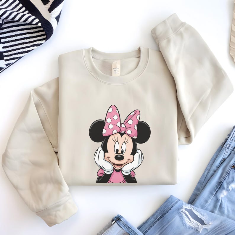 May include: A beige sweatshirt featuring a Minnie Mouse graphic. The graphic shows Minnie Mouse with a pink bow with white polka dots, a pink dress, and white gloves. The sweatshirt is folded on a white surface.