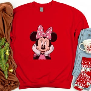 May include: A red sweatshirt with a Minnie Mouse graphic. The graphic shows Minnie Mouse with a pink bow with white polka dots. The sweatshirt is surrounded by a brown blanket, blue jeans, and a mug.