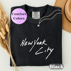 May include: A black short-sleeved t-shirt with a crew neckline featuring the white text New York City printed in a handwritten script font. The shirt has a Comfort Colors brand label at the inner neck and a silver-toned star pendant necklace.