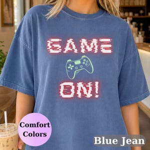 May include: A blue jean colored short-sleeve t-shirt featuring the text GAME ON! in a white, glitch-effect font with a red outline. Centered between the two lines of text is a light green graphic illustration of a video game controller.