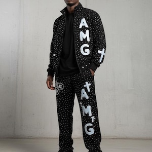 May include: Black tracksuit with white polka dots and white text that reads "AMG" and crosses. The jacket has a zip-up front and the pants have a drawstring. The outfit is worn with black shoes.