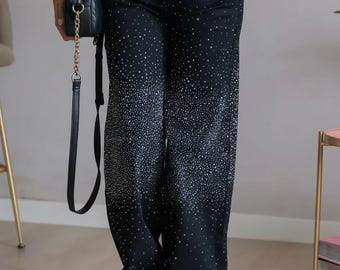 Puede incluir: Pantalones vaqueros negros de pernera ancha con un diseño brillante de pedrería que se desvanece desde la cintura hacia abajo. Los pantalones tienen un diseño clásico de cinco bolsillos y cierre de botón y cremallera. Se ve un bolso negro con una correa de cadena dorada.