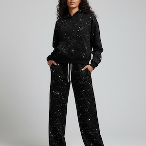 Puede incluir: Chándal negro de dos piezas con diseño de noche estrellada. El conjunto incluye una sudadera con capucha y pantalones de pernera ancha. La tela negra está cubierta de patrones blancos y plateados en forma de estrella. Los pantalones tienen un cordón blanco.