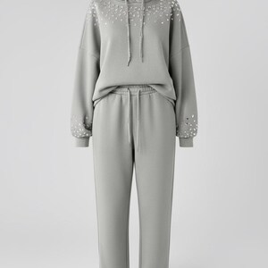 Op de afbeelding: Lichtgrijze hoodie en joggingbroek set. De hoodie heeft een trekkoord en is versierd met strass-steentjes op de schouders en mouwen. De joggingbroek heeft een taille met trekkoord. De set is gemaakt van een zacht, comfortabel materiaal.
