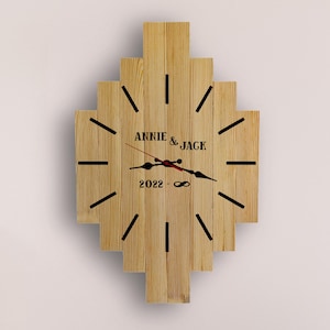 May include: Wooden wall clock with a geometric design. The clock face features the names "ANNIE & JACK" and the year "2022". Black lines mark the hours, and the clock hands are black and red.