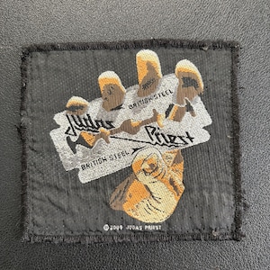 Könnte beinhalten: Schwarzer Stoffaufnäher mit dem Bandnamen "Judas Priest" und den Worten "British Steel" in einem stilisierten Design. Der Aufnäher hat einen Used-Look mit ausgefransten Kanten und einem Copyright-Hinweis.