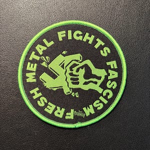 Può includere: Una toppa circolare ricamata con sfondo nero e bordo verde lime brillante. Il design presenta un pugno verde lime che spezza un simbolo di svastica, con il testo "METAL FIGHTS FASCISM" in lettere verde lime lungo il bordo.